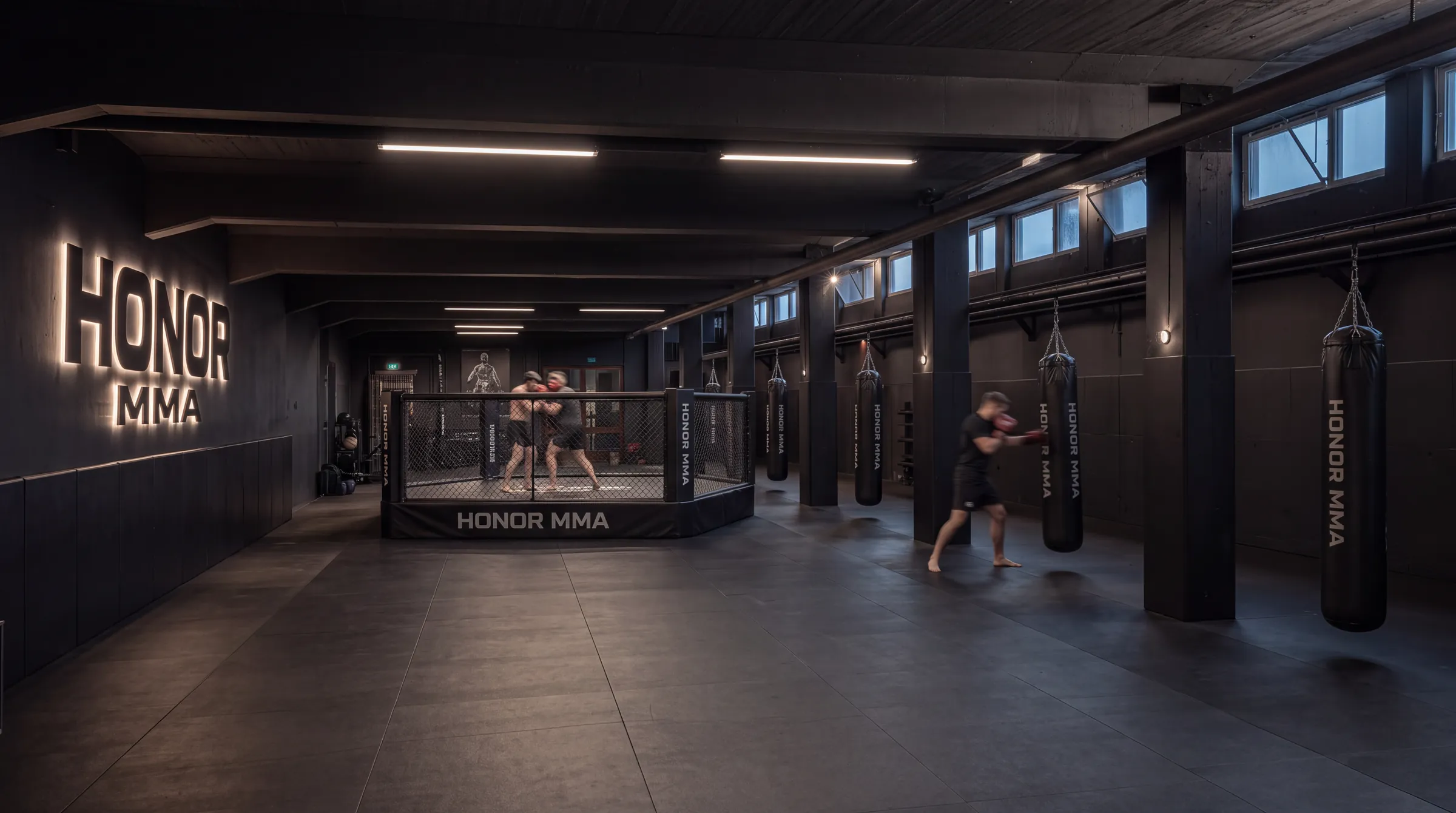 Trainingshalle der HONOR MMA Academy mit dem Octagon-Cage in der Mitte, Leuchtschrift und Sandsäcken